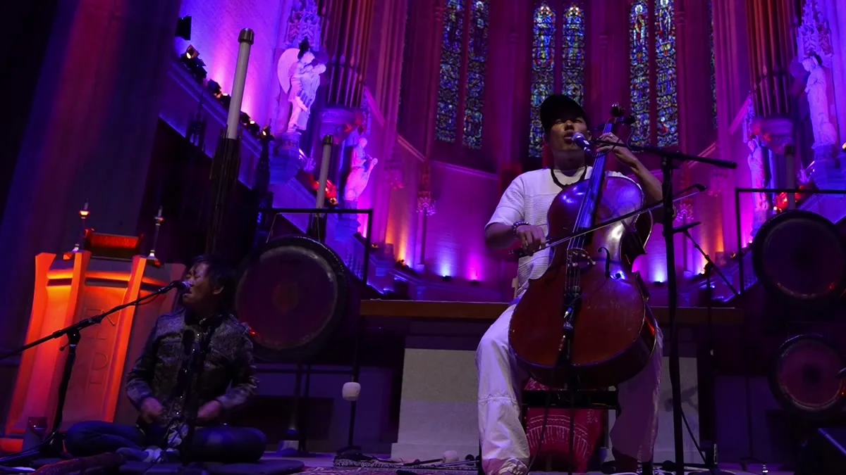 Dana Leong playing cello inside Grace Cathedral under purple light, surrounded by gongs and stained glass