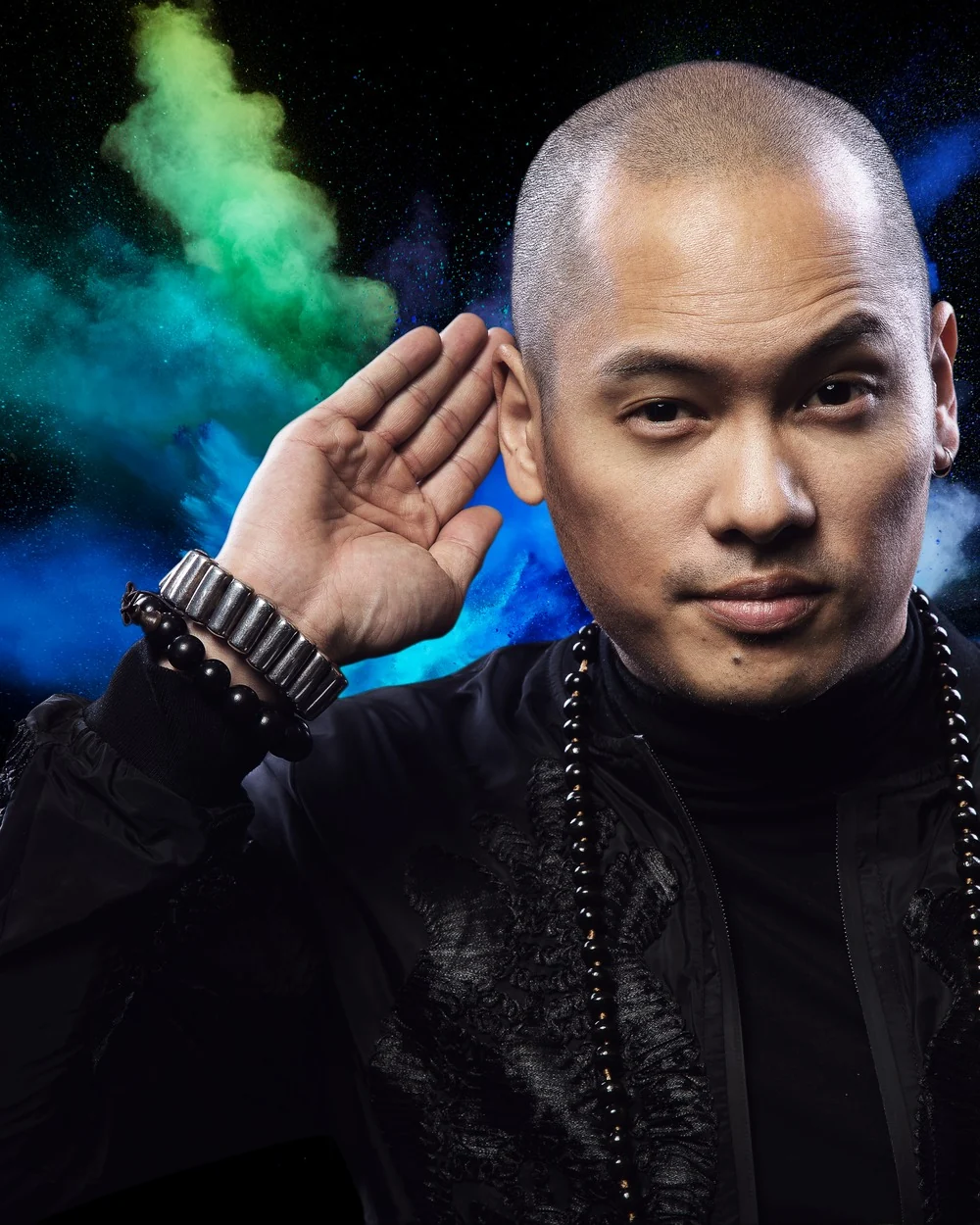 Dana Leong with a hand cupped to his ear against clouds of green and blue colour